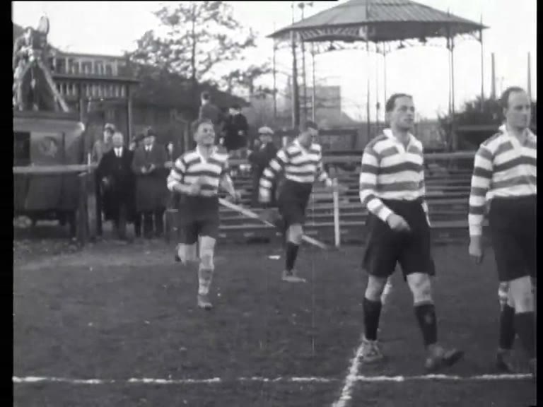 Black-and-white 1933 newsreel frame of rugby players in hooped shirts entering the field before AAC versus ARVC in Amsterdam.