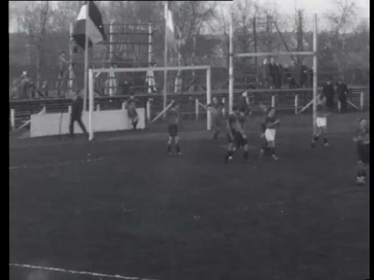 Wide black-and-white 1933 newsreel frame of AAC and AVRC playing rugby near the posts at an Amsterdam ground.