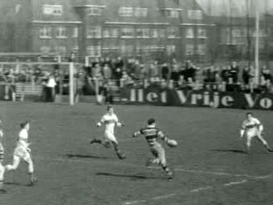 Black-and-white newsreel frame of an AAC rugby player in a broad-banded shirt carrying the ball towards three Bank of England defenders in Amsterdam in April 1955.