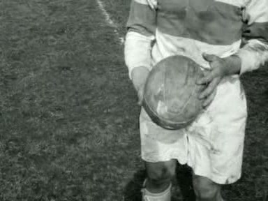 Black-and-white close view of an AAC rugby player in a broad-banded long-sleeved shirt holding a worn leather rugby ball in Amsterdam in April 1955.