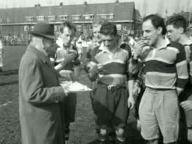 Black-and-white newsreel frame of five AAC rugby players in broad-banded shirts taking refreshments beside the pitch in Amsterdam in April 1955.