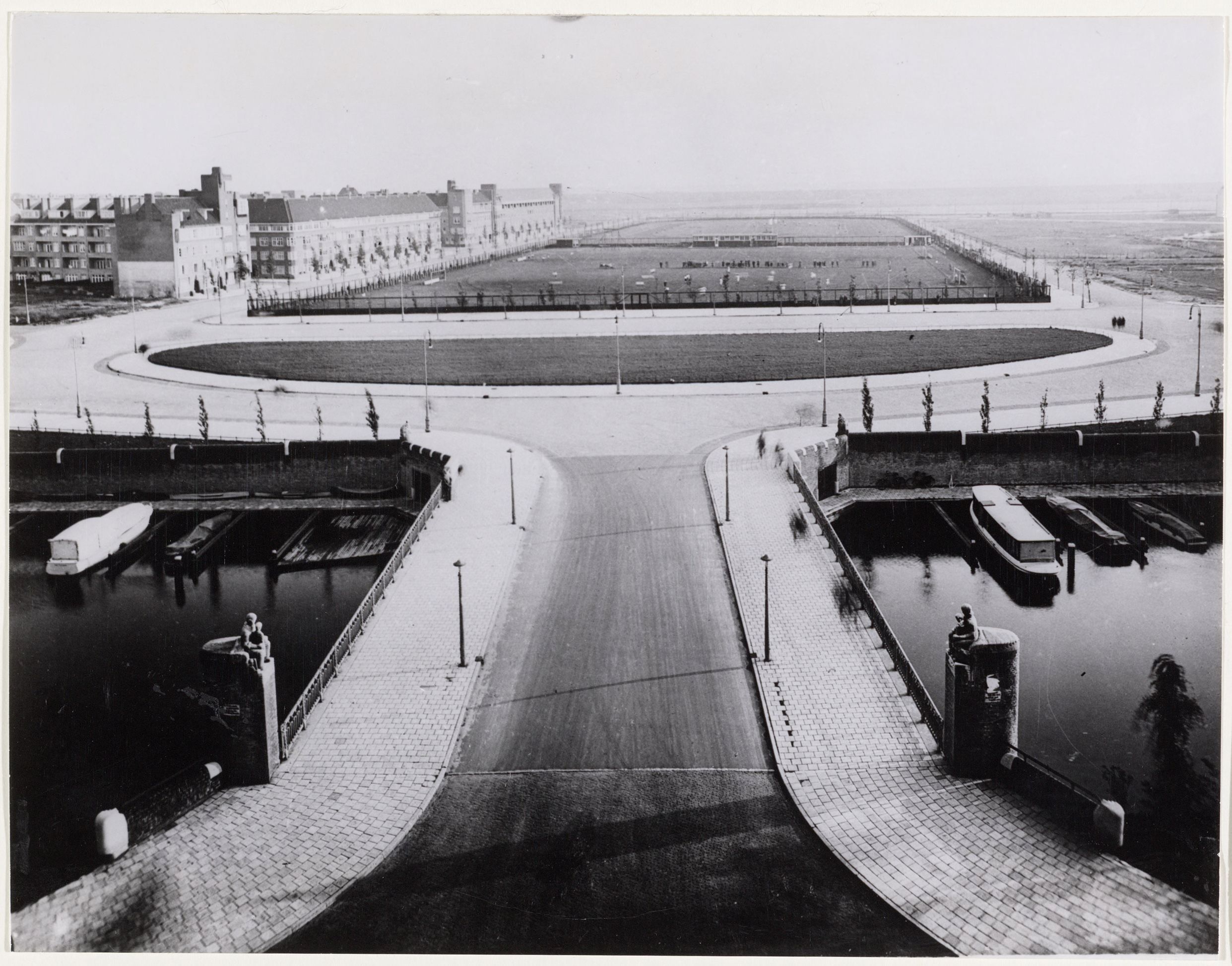 Black-and-white view across a bridge towards the Olympiaplein sports ground in May 1932, with an athletics track and central field beyond the road.