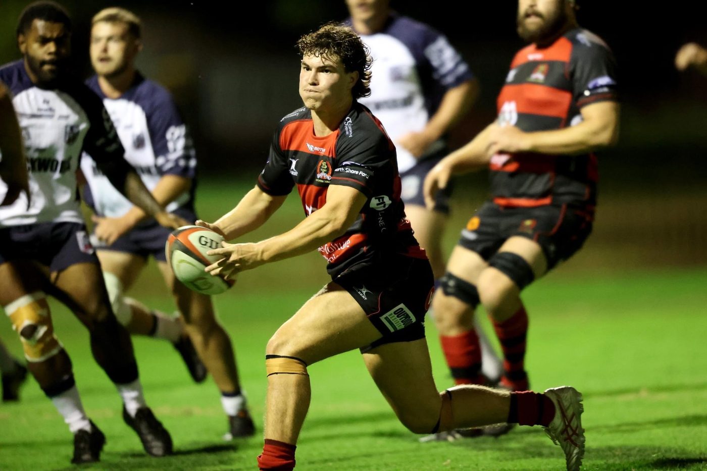 Angus James carries the ball to the line under floodlights in red-and-black hooped club kit