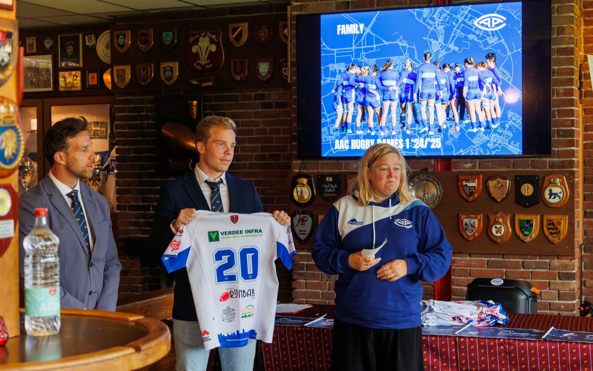 AAC jersey with sponsor branding presented in the clubhouse beside a screen showing the club crest and squad