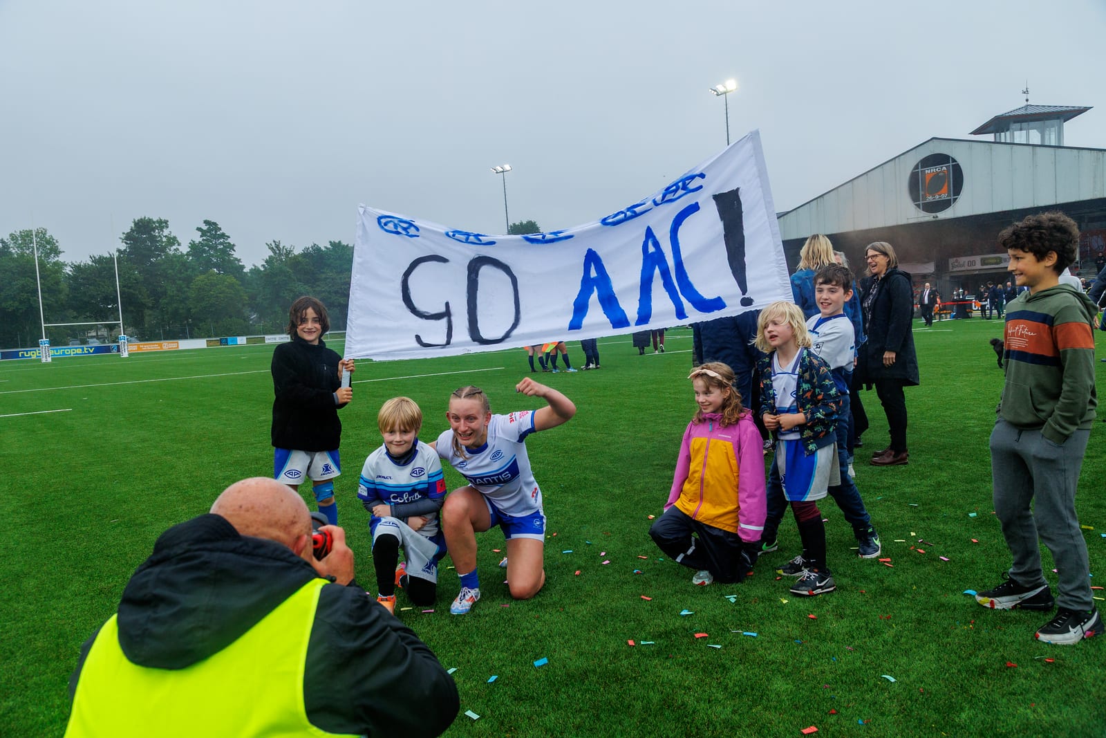 AAC player kneeling with cubs holding a "Go AAC!" banner on the pitch after full-time